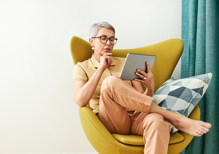 Thoughtful mature woman with closed eyes sitting at home holding a digital tablet