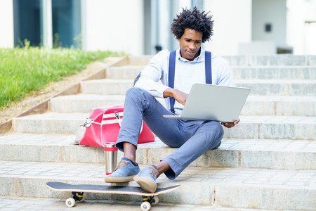 Black businessman with afro hair and skateboard using his laptop computer sitting on some steps