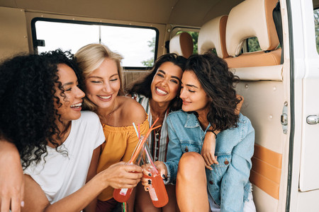 Two smiling women toasting with bottles in van  Close friends enjoying summer road trip