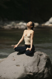 Serene beautiful young woman meditating in lotus pose on sunny rock