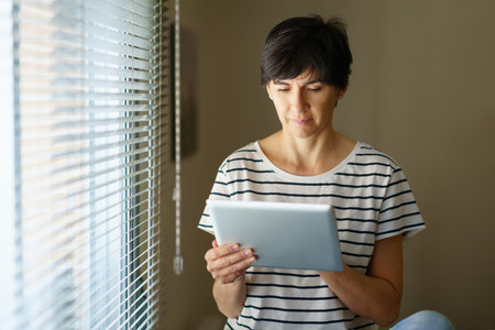Middle aged woman consulting on her digital tablet sitting near the window at home