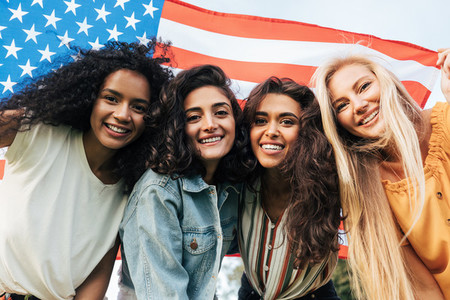 Four diverse women friends looking at the camera under the American flag  Cheerful females celebrating the 4th of July outdoors