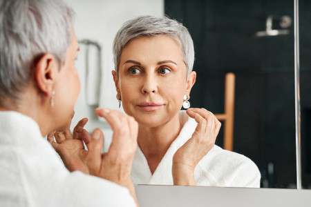 Smiling mature woman looking at her reflection while applying moisturizer on the face
