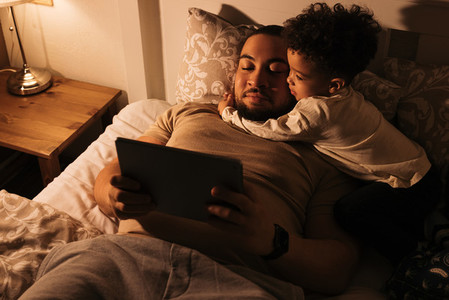Little boy embracing his father while them lying on a bed Young father reading book to his child from a digital tablet before sleeping