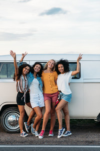 Four female friends in casuals standing at minivan on road raised hands up
