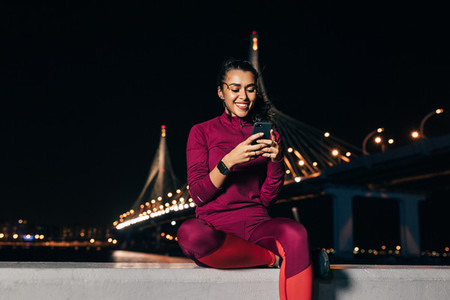 Smiling sportswoman sitting on an embankment using smartphone at night