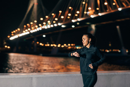 Young fit woman in sport clothes looking at smartwatch while jogging on an embankment at night