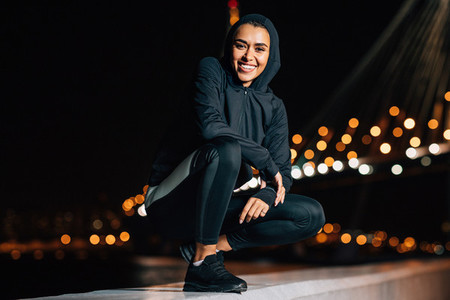 Cheerful middle east woman in sport hoodie shirt sitting outdoors at night looking at camera