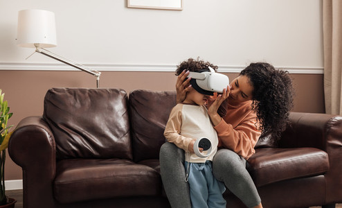 Young mother sitting on couch helping her son wear VR goggles