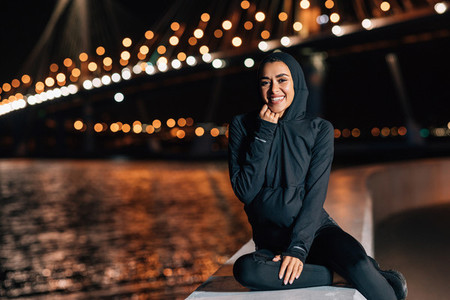 Beautiful middle east woman in hoodie sitting on embankment at night and resting after workout