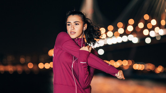 Young fitness woman in headphones stretching her arm at night during a workout