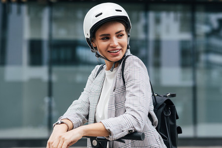 Young businesswoman with electric scooter looking away Cheerful female in safety helmet wearing backpack standing outdoors