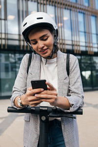 Young woman with a safety helmet leaning on an electrical scooter typing on her cell phone