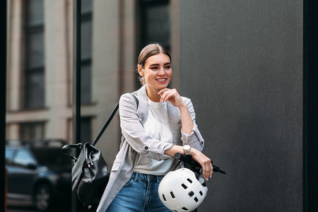 Smiling confident woman with an electrical scooter safety helmet and backpack standing at the building