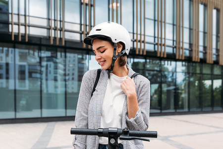 Beautiful smiling woman wearing helmet standing outdoors with an electric scooter in front of building