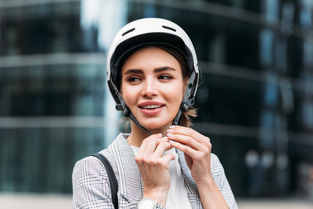 Smiling businesswoman strapping on a cycling helmet while standing in the city Young female putting a white helmet on her head