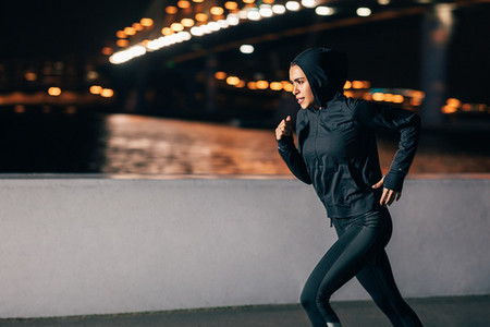 Young female athlete in hoodie sprinting at night in the city