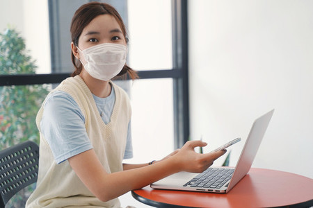 Young asian millennial woman wearing mask and using laptop and s
