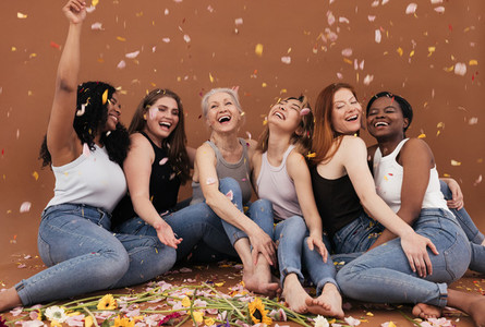 Group of happy women sitting on the brown background while petals from flowers falling  Laughing females of different races and ages under petals from flowers