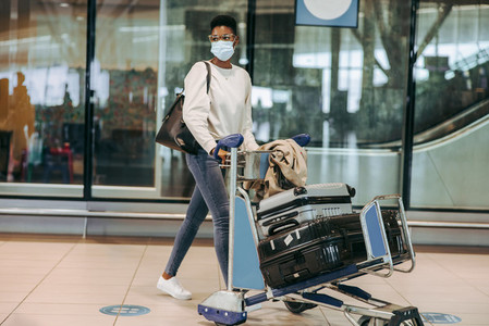 Woman traveler in face mask with luggage trolley at airport