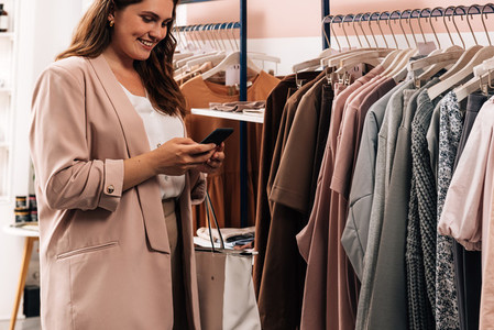 Smiling woman using cell phone while standing at a rack in a clothing store  Plus size female holding smartphone while shopping