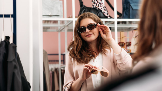 Smiling woman trying fashionable sunglasses in front of a mirror  Buyer choosing eyewear in a boutique