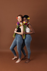 Two women with different skin color posing against a brown background  Cheerful females with flowers standing together looking at camera