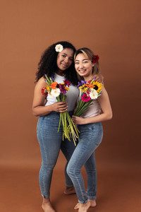 Asian and mixed race women posing against a brown background  Two smiling females with bouquets looking at camera