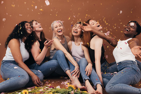 Group of six laughing women of different ages sitting under falling flower petals  Multi ethnic smiling females having fun in studio while sitting on brown background