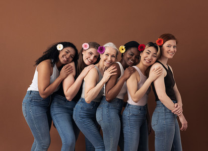 Group of six cheerful women with flowers in hair stand behind each other  Smiling females of different ages posing against a brown background