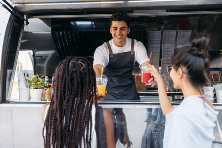 Back view of two females buying drinks from a male owner at a food truck  Smiling entrepreneur giving drinks to clients