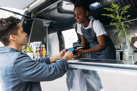 Young man with credit card paying to a saleswoman at a food truck  Female entrepreneur in apron receiving payment from a customer