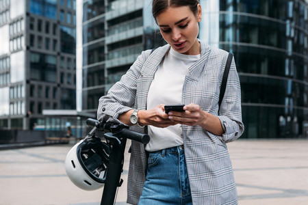 Young woman in stylish clothes holding smartphone while standing in front of an office building