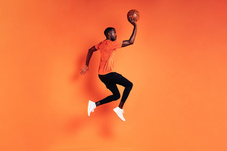 Male athlete jumping with basket ball Side view of sportsman exercising over orange background