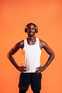 Happy guy wearing wireless headphones standing against an orange background with hands on his waist