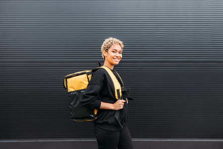 Side view of a smiling delivery girl with backpack standing at black wall outdoors