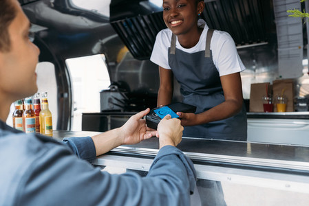 Customer paying with a credit card at a food truck  Smiling saleswoman in apron holding a pos terminal while buyer making NFC transaction