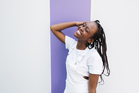 Happy woman with closed eyes wearing white clothes leaning on white wall with purple stripe
