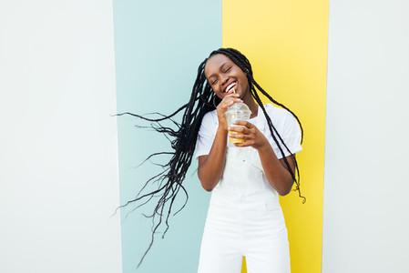 Happy woman with long braided hair having fun while drinking orange juice at a wall with blue and yellow stripes