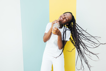 Cheerful woman with closed eyes drinking orange juice while dancing at a wall with blue and yellow stripes