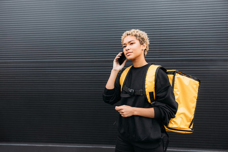 Portrait of a delivery girl with yellow thermal backpack on her back talking on smartphone at a black wall