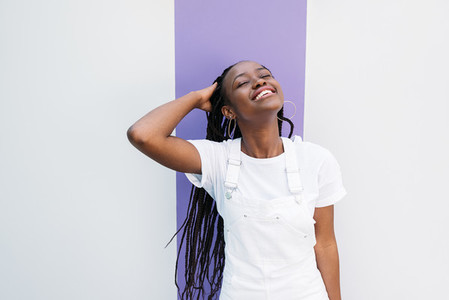 Young smiling woman in casuals adjusting hair with closed eyes against white wall with purple stripe