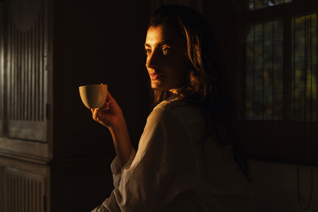 Carefree young woman having a cup of coffee in a dark hotel room