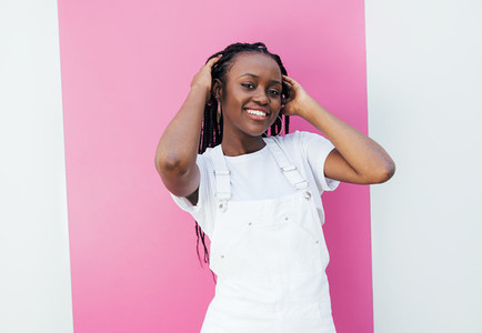 Smiling woman with braids adjusting hair while standing at white wall with pink stripe