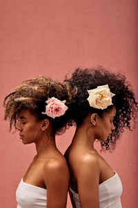 Two women with flowers in their hairs standing back to back at a pink wall with closed eyes