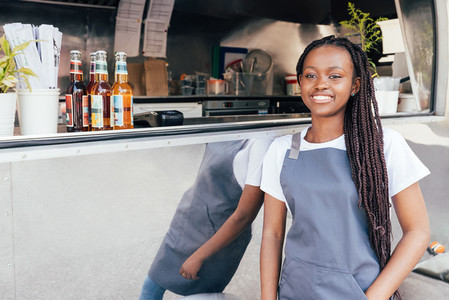 Smiling waitress with long braids leaning on food truck looking away