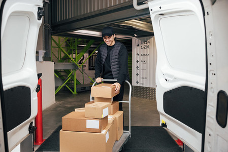 Smiling courier in uniform checking cardboard boxes on a cart while standing in a warehouse