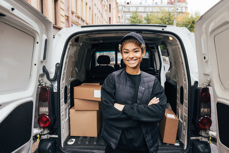 Portrait of a smiling delivery girl wearing a cap and uniform standing with crossed arms at van trunk