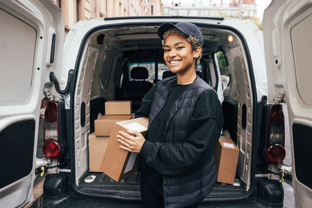 Portrait of a happy woman courier holding a cardboard box while standing at car trunk