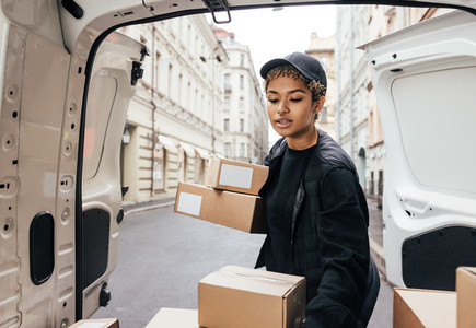 Woman courier in uniform carefully unloading cardboard boxes from car for delivery
